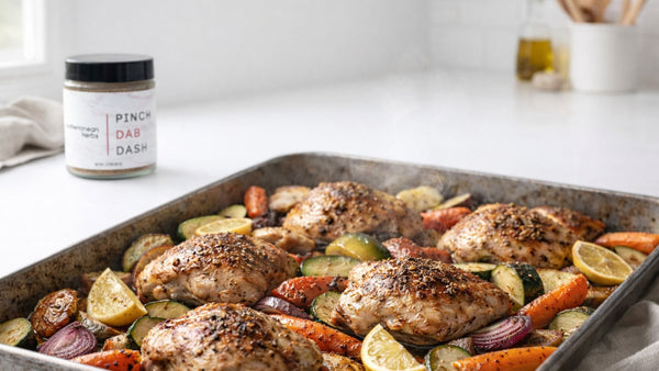Roasted chicken and vegetables on a baking tray with a container of seasoning in the background.