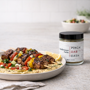 Plated dish of skewered meat with vegetables and a jar of 'Pinch Dab Dash' seasoning on a light surface.