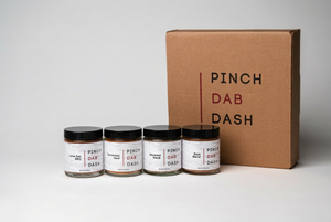 Four jars of 'Pinch Dab Dash' on a white surface with a cardboard box in the background.